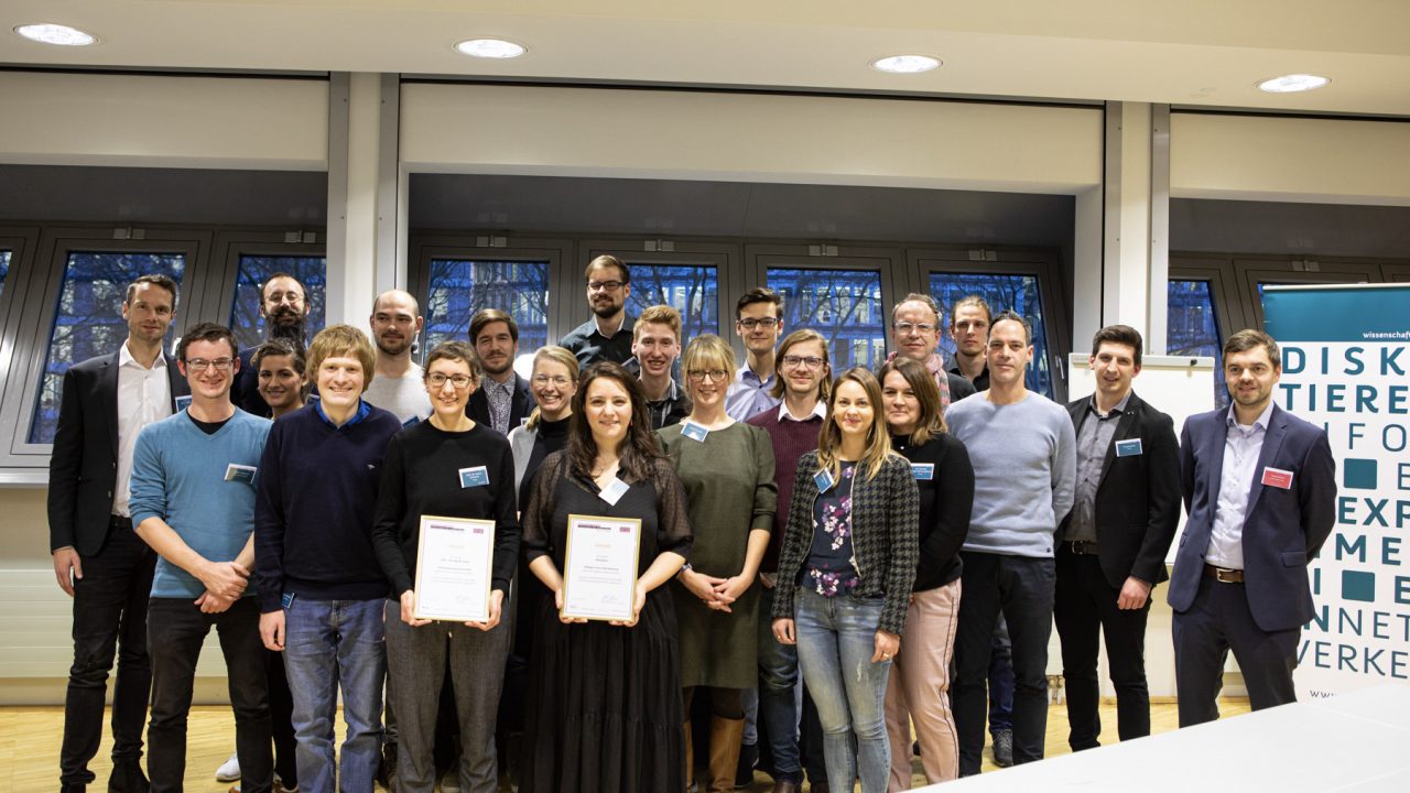 Eine Gruppe von 24 Personen steht in einem Raum und lächelt in die Kamera. Im Hintergrund hängen Banner mit dem Logo von Wissenschaft im Dialog. Zwei der abgebildeten Gewinner*innen des Hochschulwettbewerbs halten eingerahmte Urkunden in den Händen und präsentieren sie stolz.