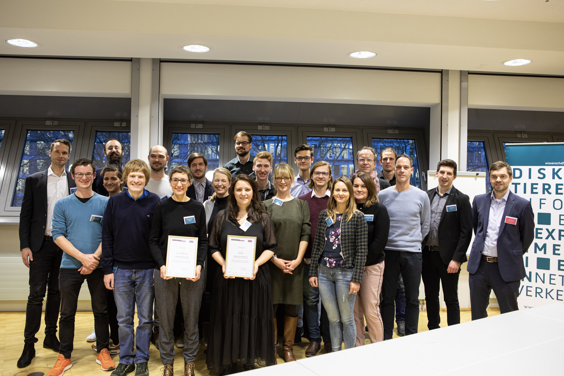 Eine Gruppe von 24 Personen steht in einem Raum und lächelt in die Kamera. Im Hintergrund hängen Banner mit dem Logo von Wissenschaft im Dialog. Zwei der abgebildeten Gewinner*innen des Hochschulwettbewerbs halten eingerahmte Urkunden in den Händen und präsentieren sie stolz.